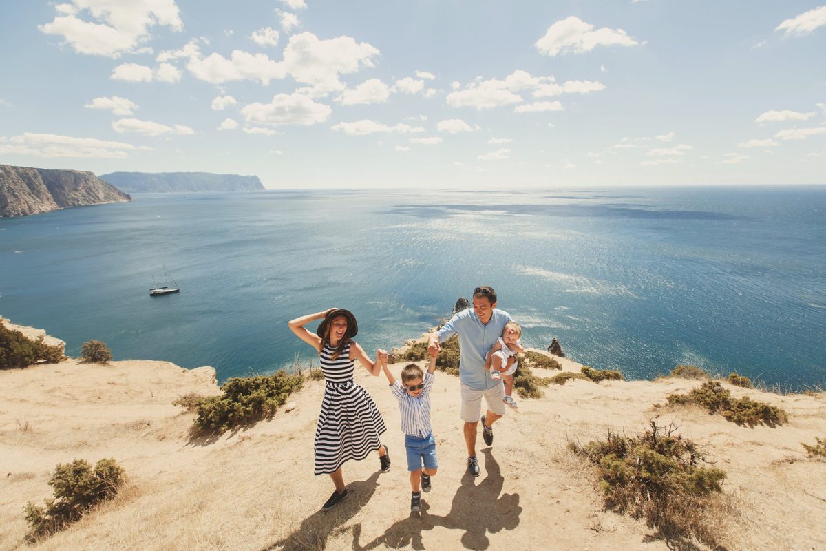 Une famille joyeuse, parents, un petit garçon et un bébé, marche main dans la main sur une falaise face à la mer bleue