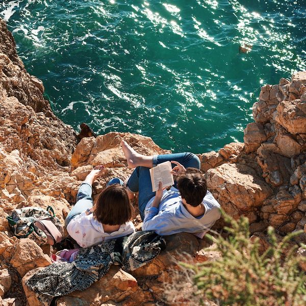 Un couple allongé sur des rochers au bord d’une mer turquoise, un livre à la main